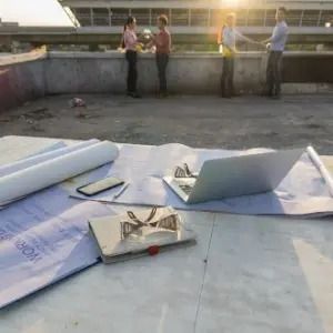 Rooftop with rolled plans, laptop, and tools in foreground; three people stand by a low wall in the background.