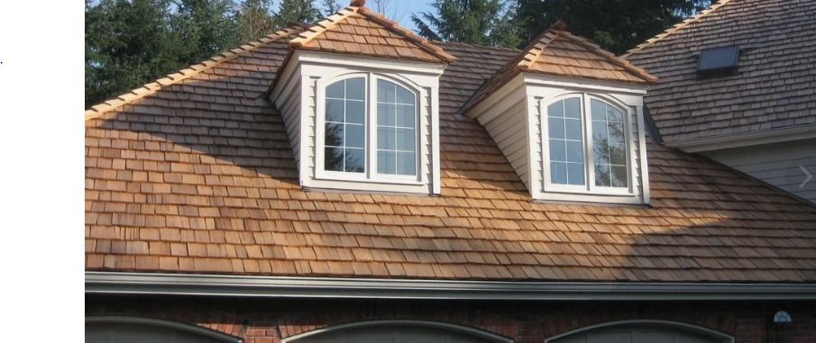 A brown shingled roof with two white dormer windows in sunlight