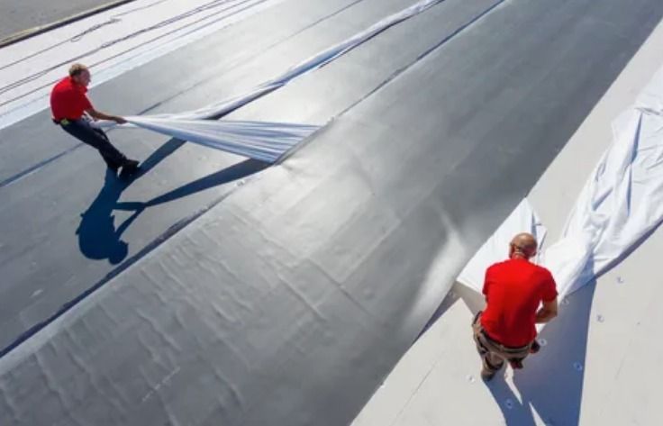 Two workers in red shirts stretching a large gray tarp across a rooftop
