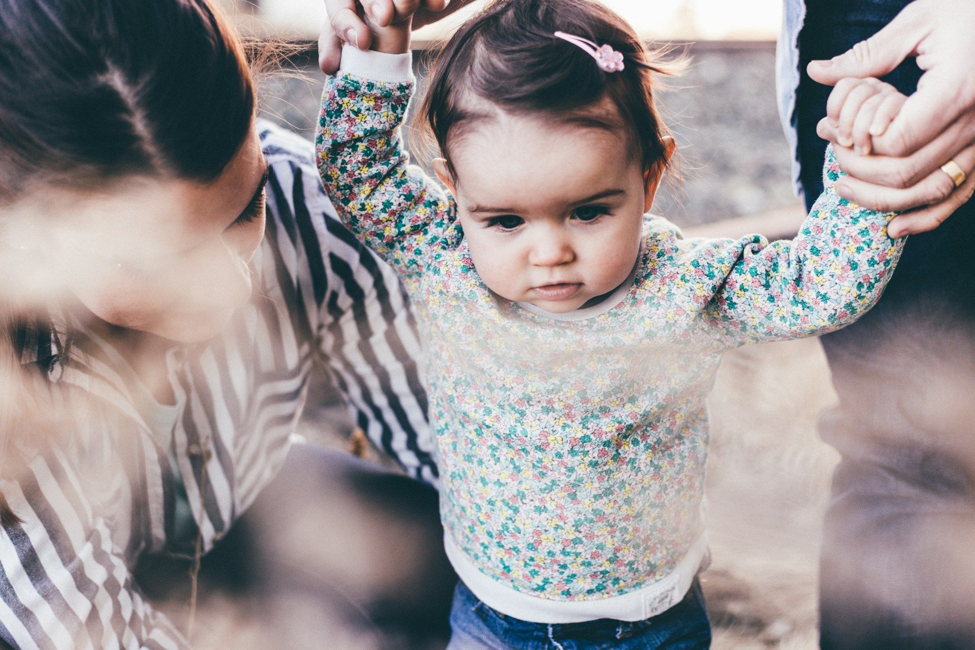 Parents helping a toddler walk outside; child looks focused, hands raised, surrounded by greenery.
