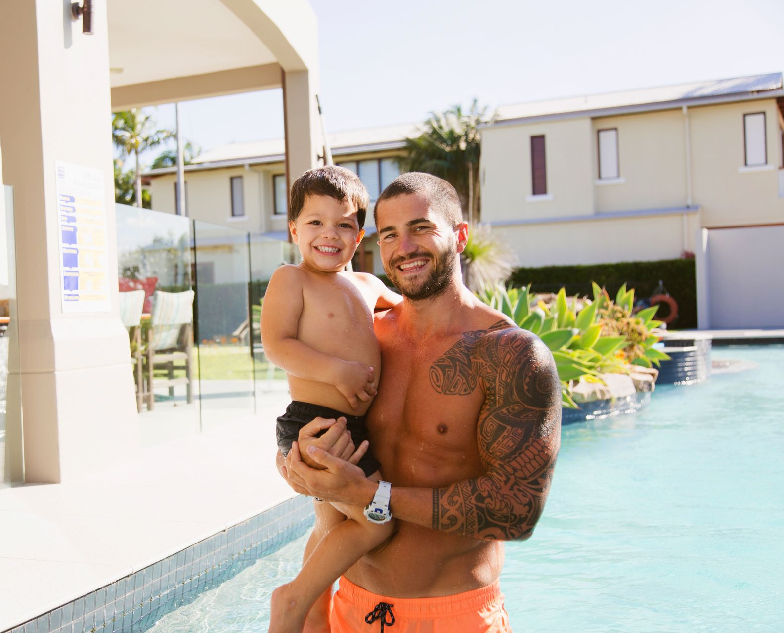 Man holding a child, both smiling, by a pool. Man has tattoos. Sunny outdoor setting.