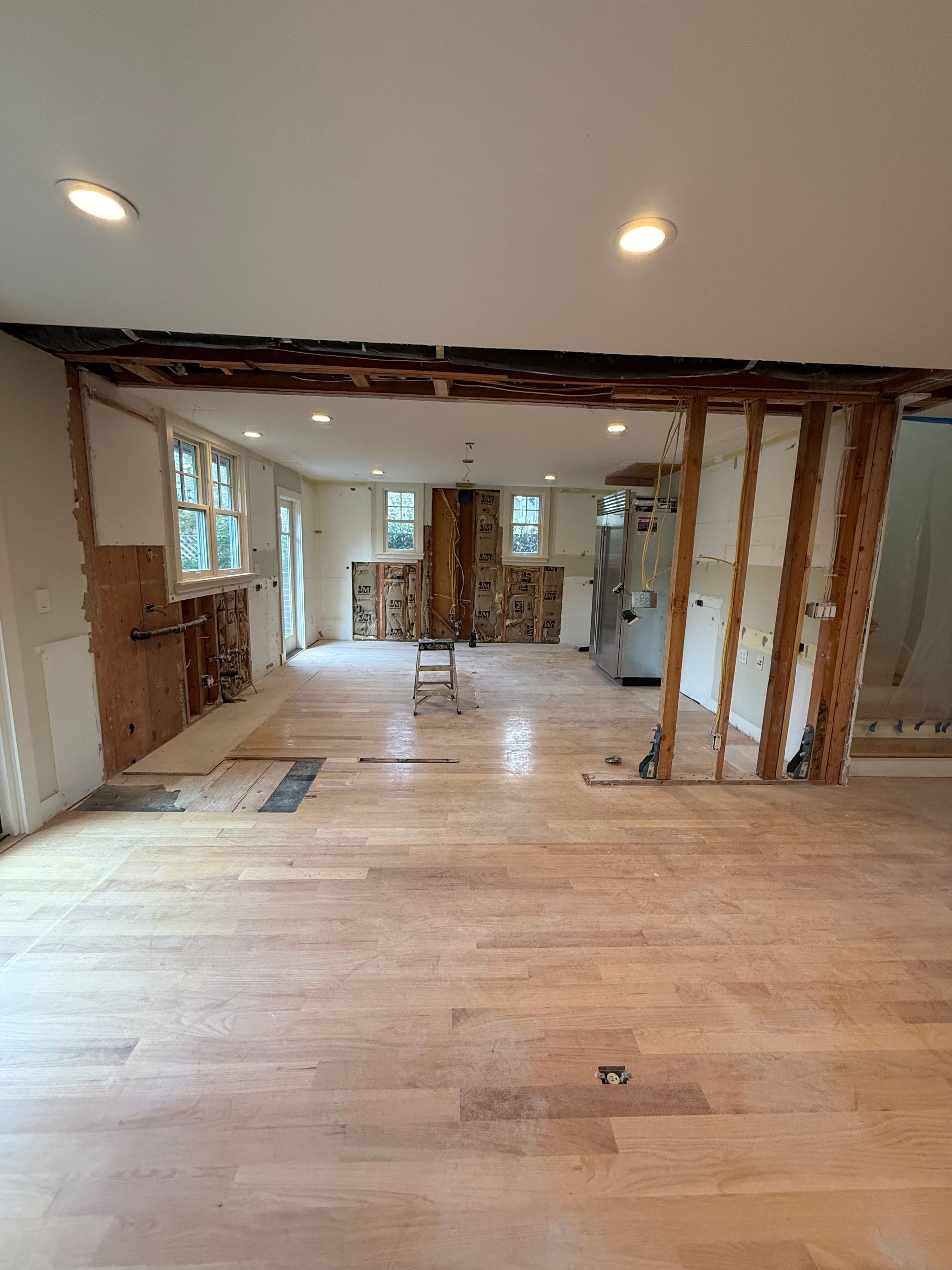 A room under renovation with wooden ceiling beams, drywall patches, ladders, and scaffolding on a hardwood floor.