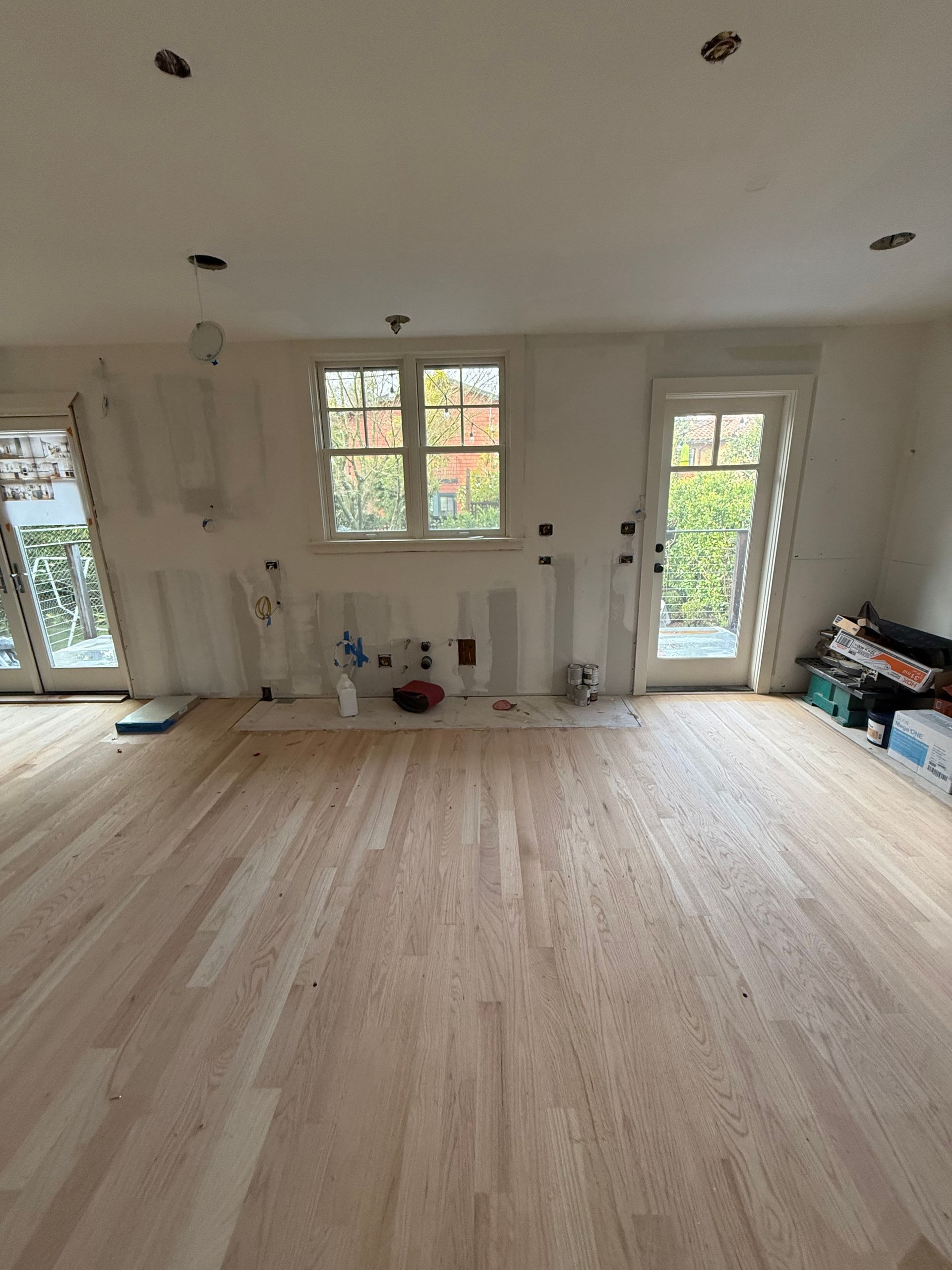 A room under renovation with wooden ceiling beams, drywall patches, ladders, and scaffolding on a hardwood floor.