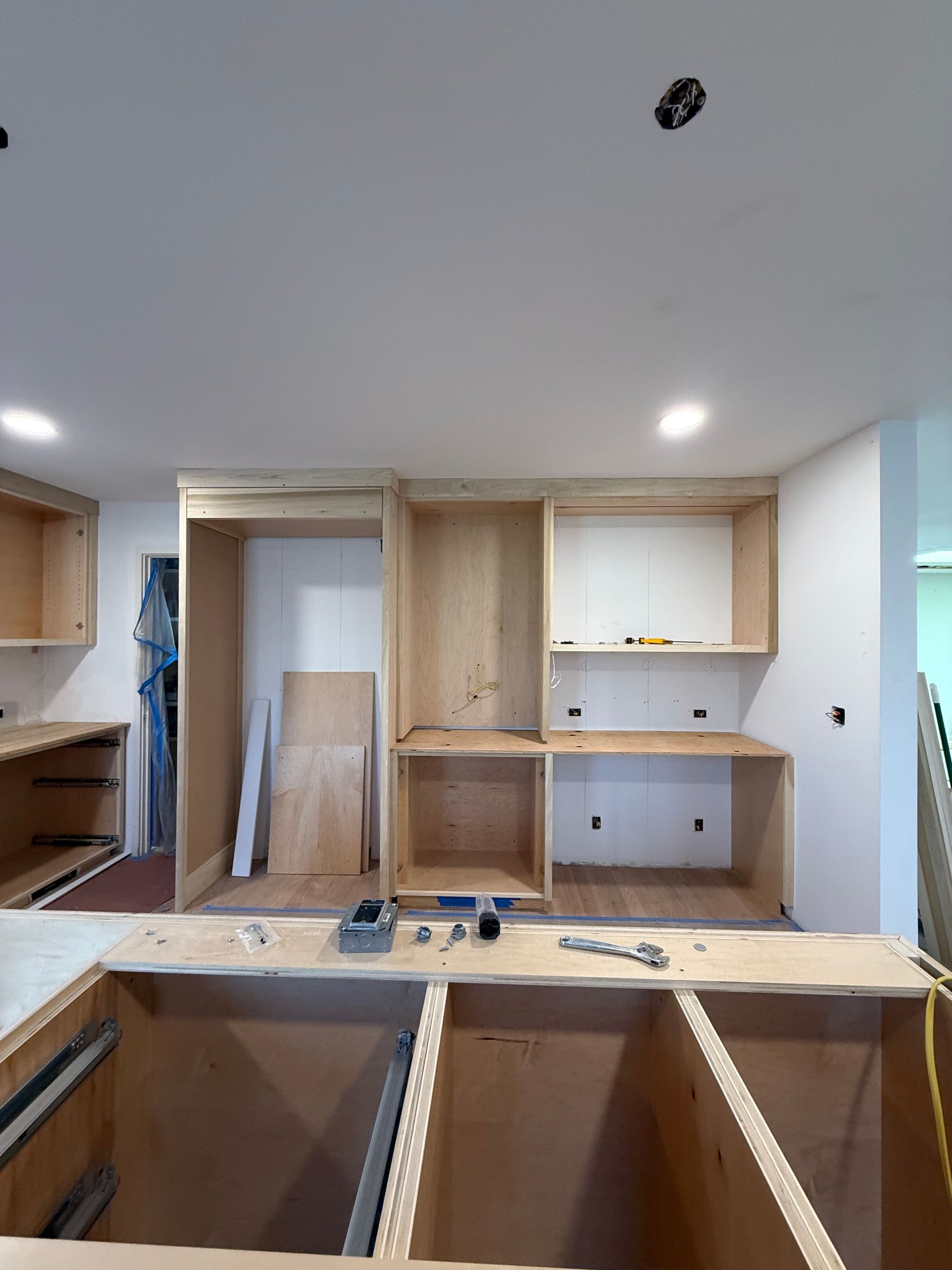 A room under renovation with wooden ceiling beams, drywall patches, ladders, and scaffolding on a hardwood floor.