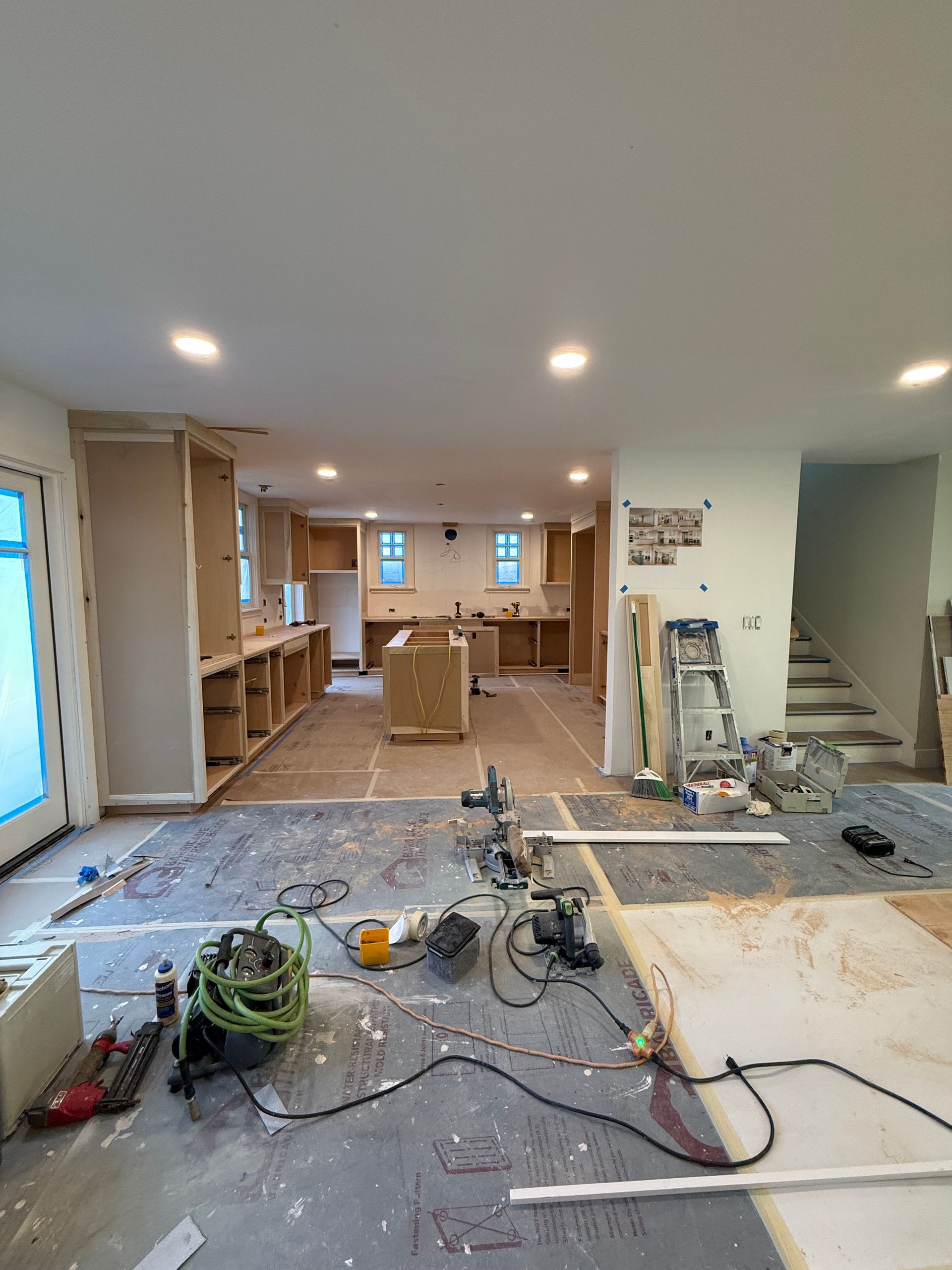 A room under renovation with wooden ceiling beams, drywall patches, ladders, and scaffolding on a hardwood floor.