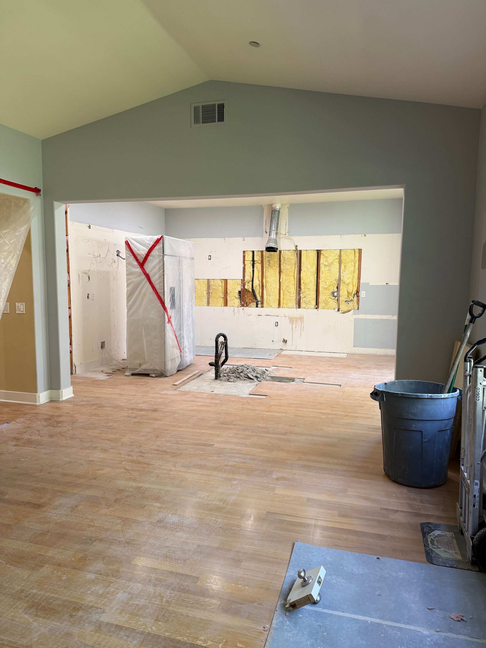 A room undergoing interior renovation with exposed wall studs, plastic sheeting, a grey trash bin, and wood flooring.