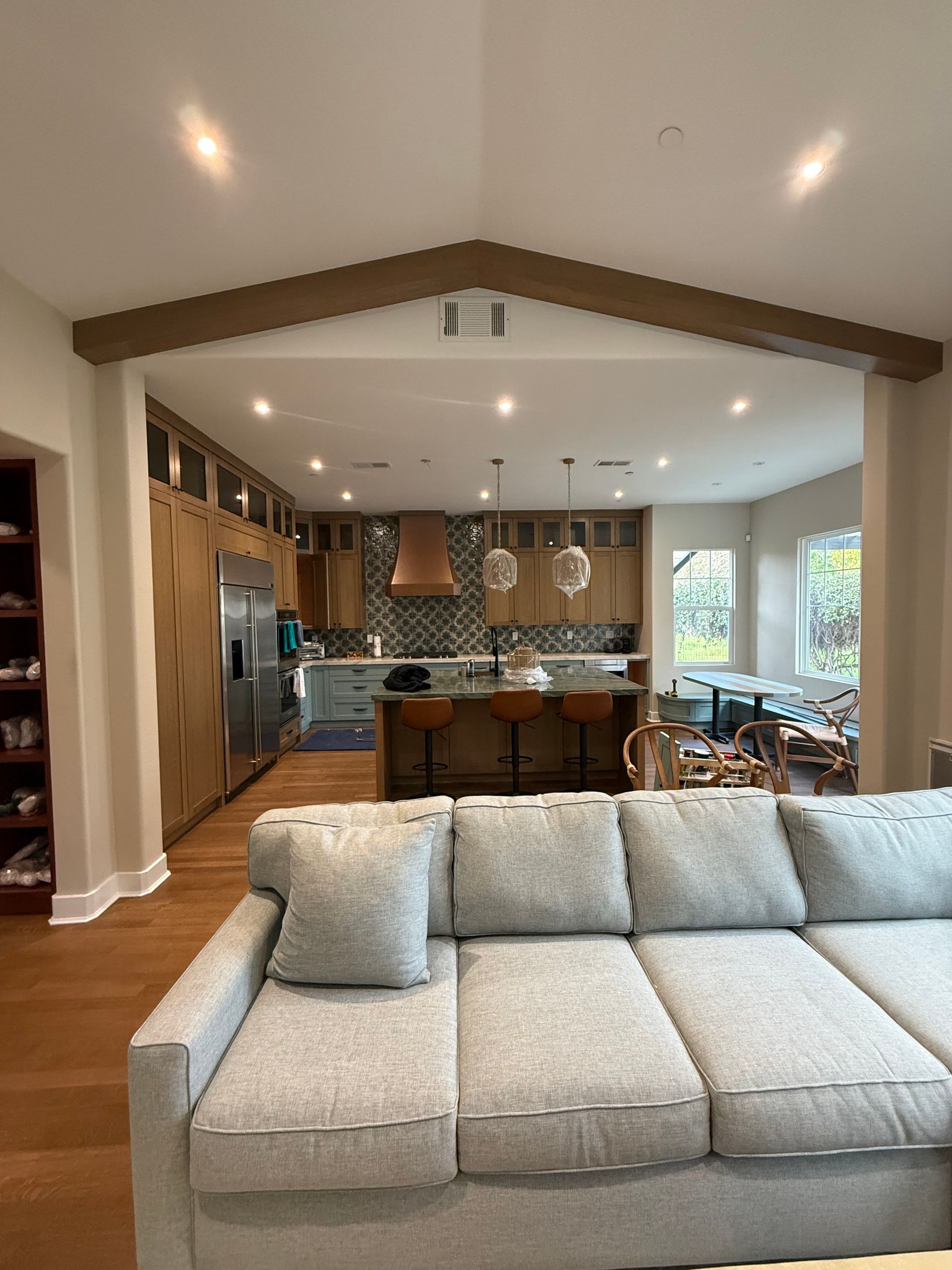A modern living room and open kitchen featuring a light gray sofa in the foreground and wooden cabinetry in the back.