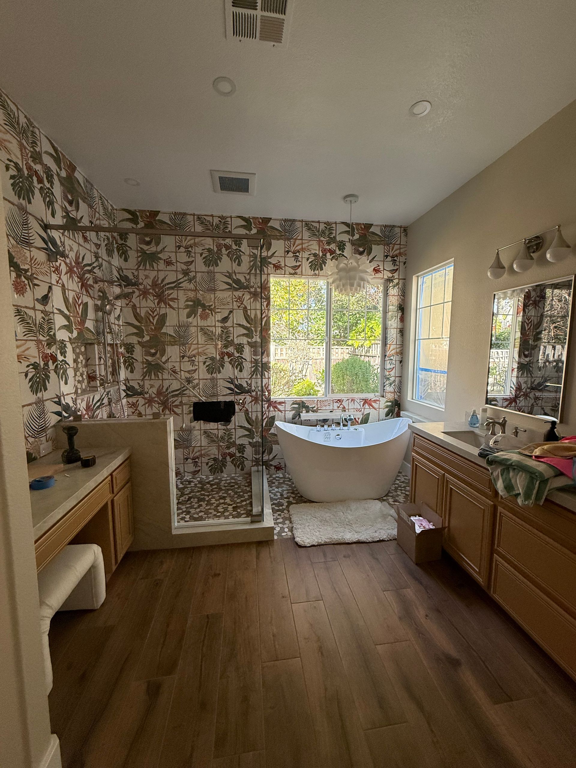 Bathroom with a freestanding tub, wood-toned vanity, and floral wallpaper behind a shower area with dark-framed glass.