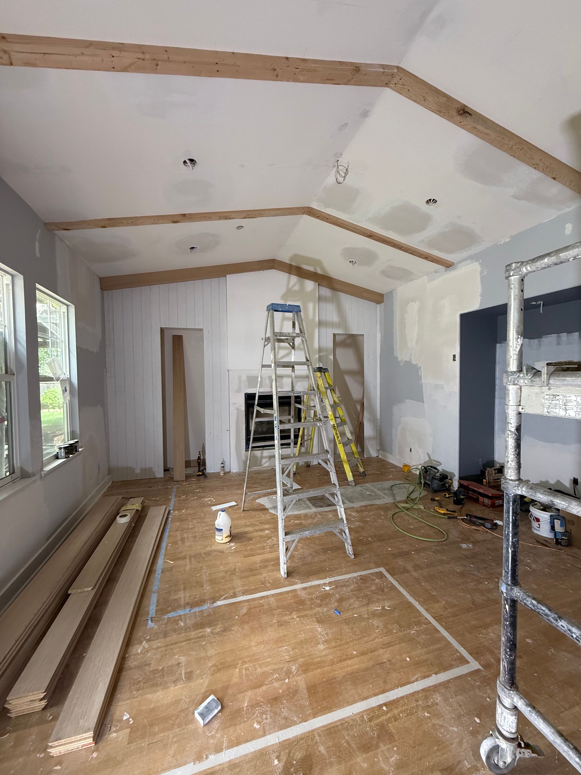 A room under renovation with wooden ceiling beams, drywall patches, ladders, and scaffolding on a hardwood floor.