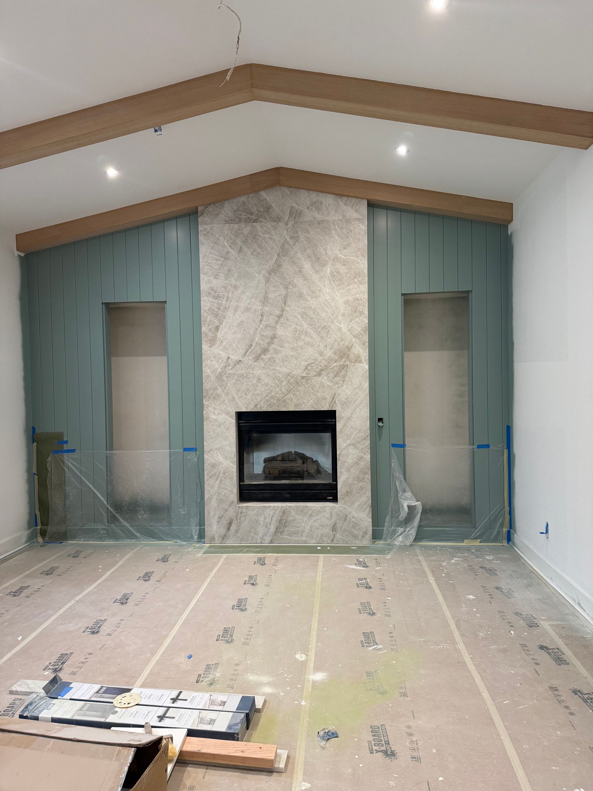 A fireplace with a marble facade and two recessed wall niches, framed by wooden ceiling beams in a room under construction.