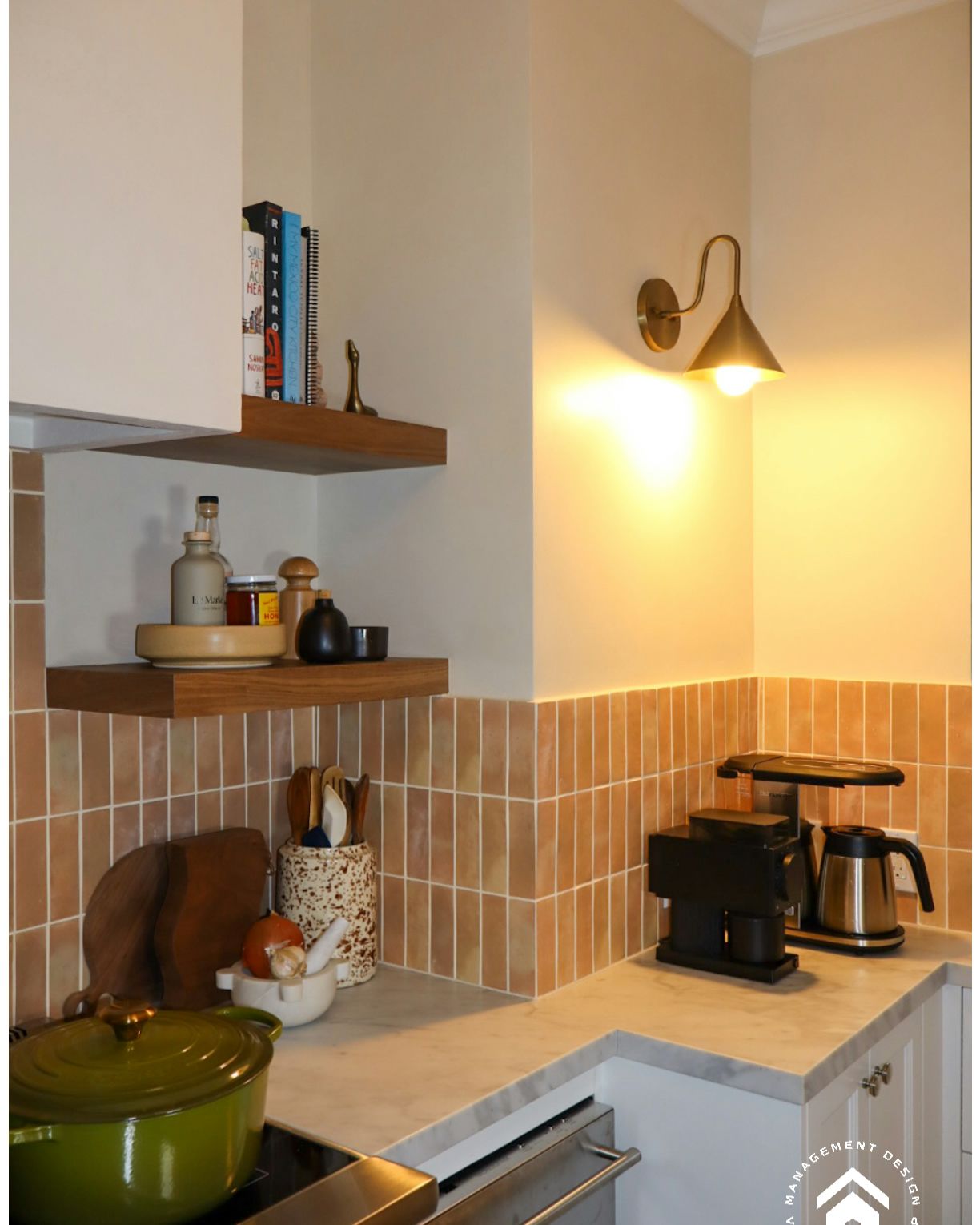 A kitchen counter with wood shelves holding books and jars, a wall sconce, coffee maker, and a green pot on the stove.