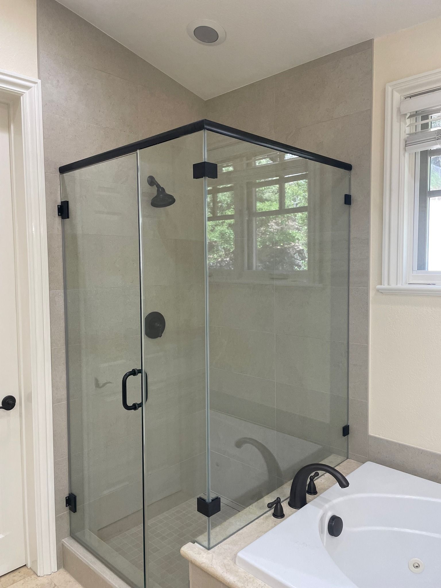 A frameless glass shower enclosure with black hardware, situated in a tiled bathroom corner next to a white bathtub.