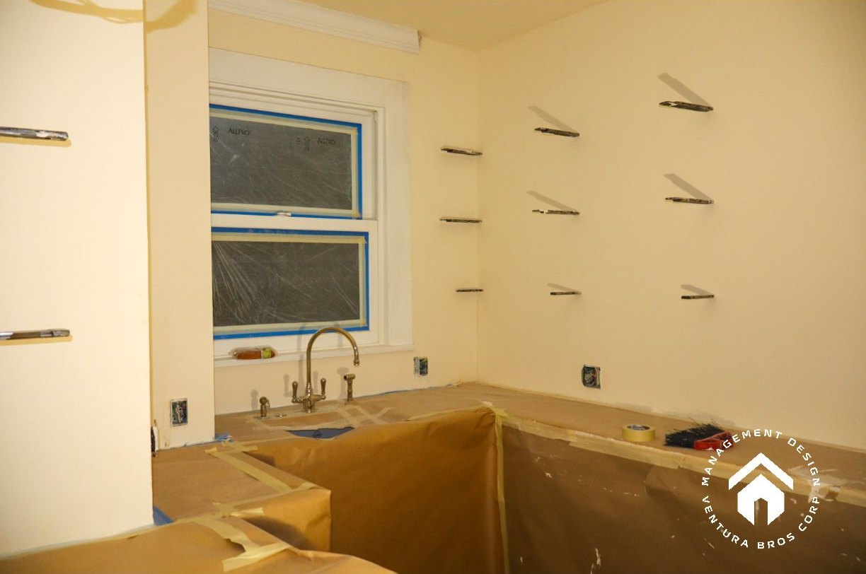 An unfinished kitchen renovation with light yellow walls, a window above the sink, and floating shelf brackets mounted.