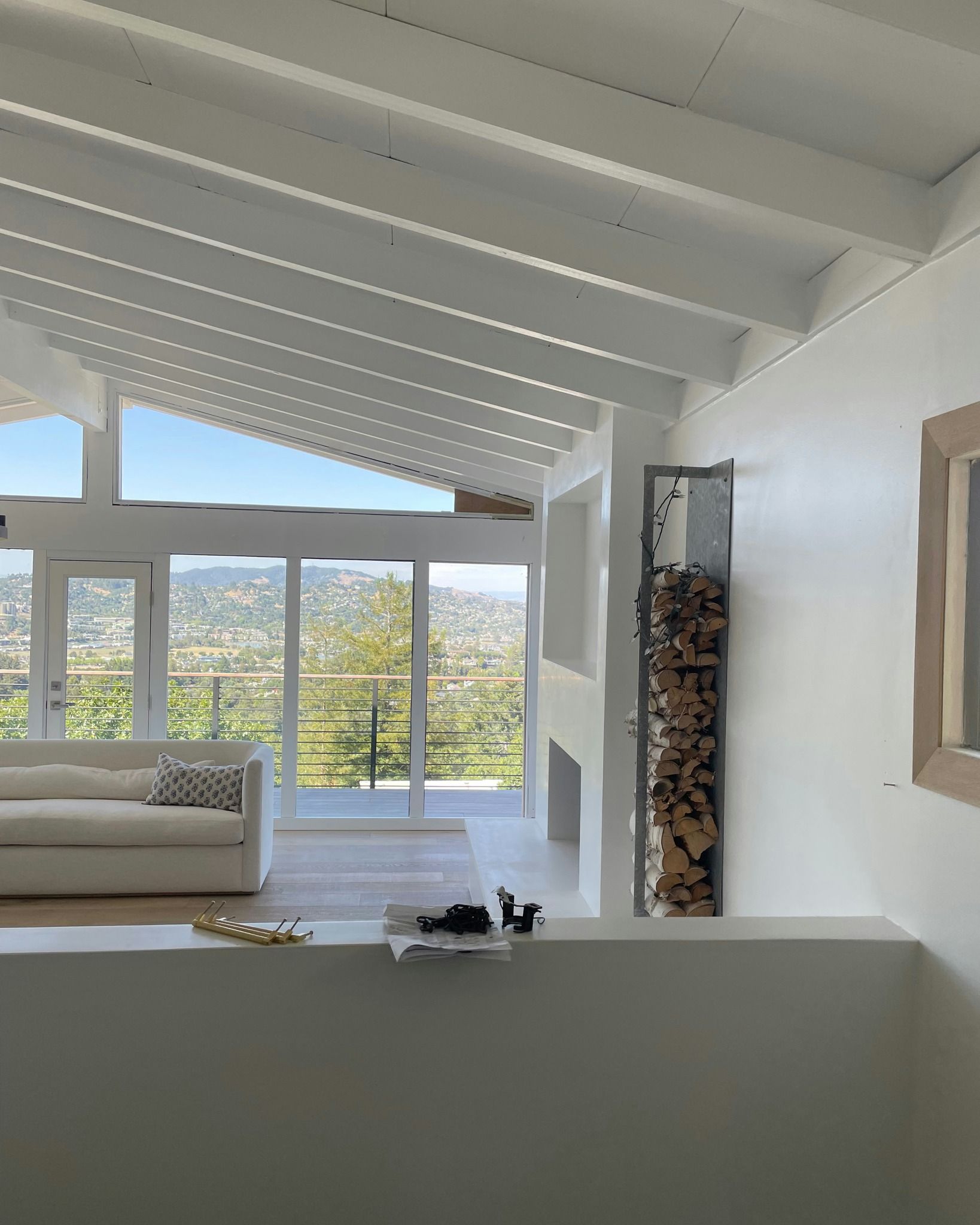 A bright, modern living room with white vaulted ceilings, large windows overlooking a valley, and a vertical wood rack.