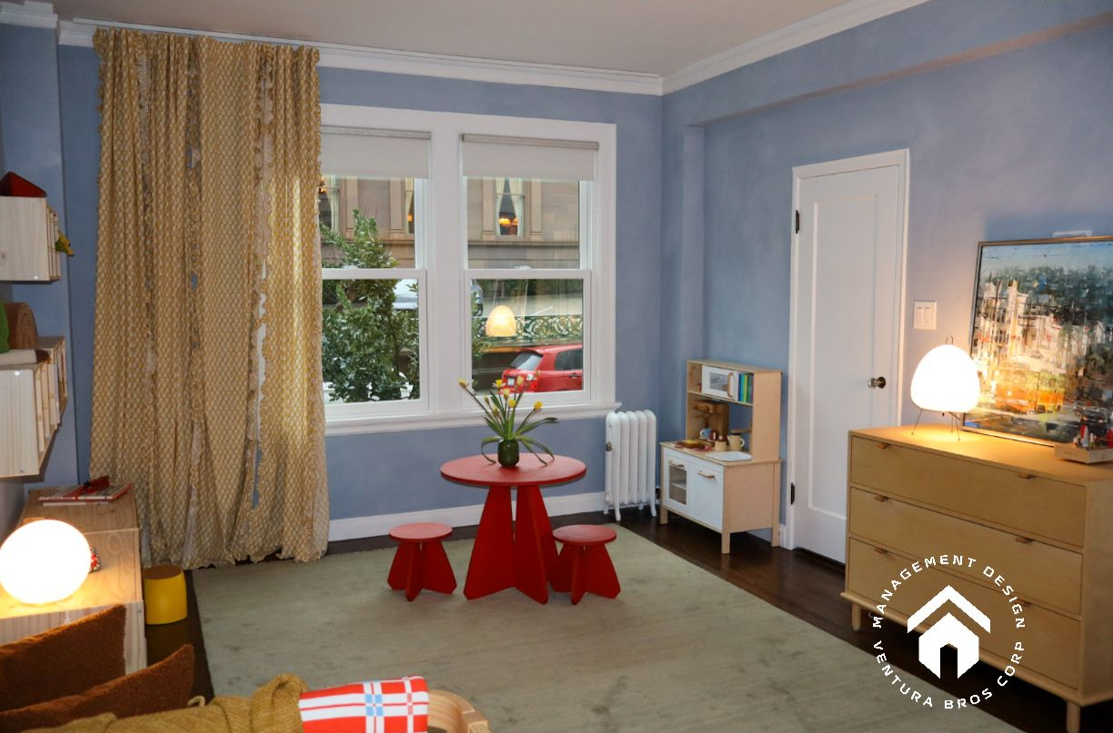 A blue nursery with a red table, two small red stools, a toy kitchen, a dresser, and patterned curtains over windows.