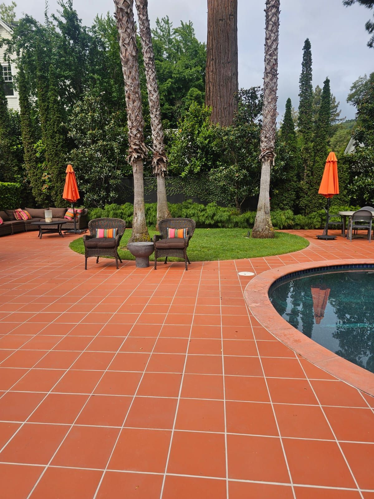 Poolside patio with terracotta tiles, lounge chairs, orange umbrellas, and tall trees beside a curved pool