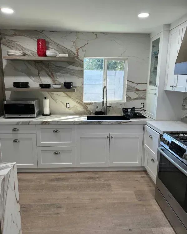 Modern kitchen with white cabinets, light wood floors, and a marble-patterned backsplash wall with two floating shelves.
