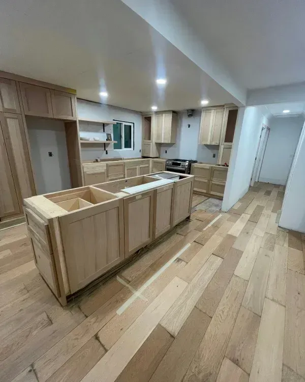 A kitchen under renovation with unfinished light wood cabinets, an island, and light wood flooring.