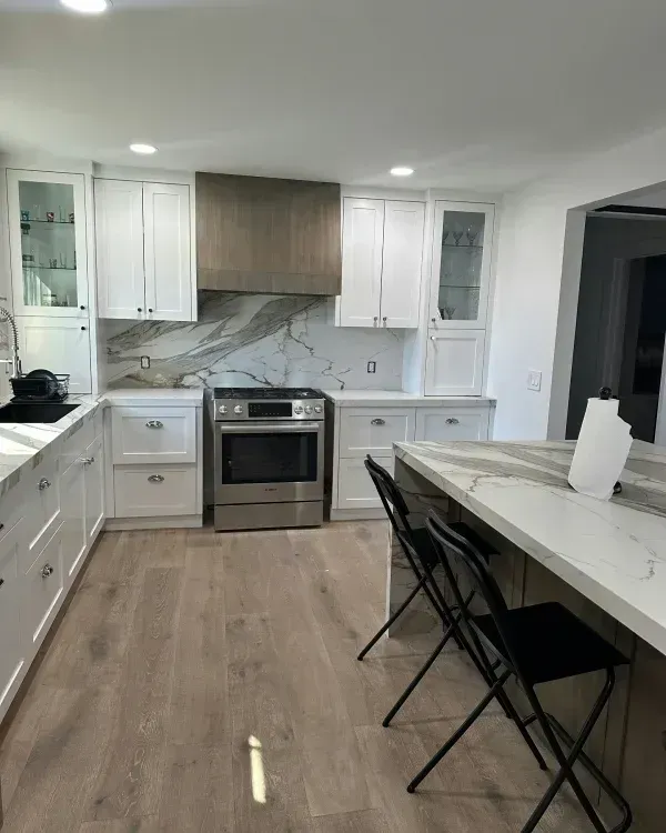 Modern white kitchen with wood-toned range hood, marble-style backsplash, hardwood floors, and island with bar stools.