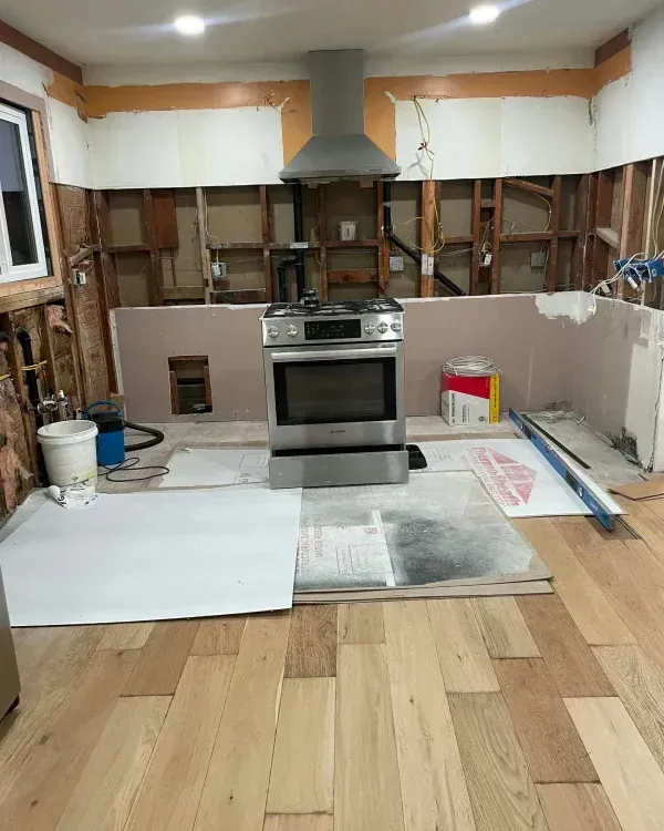 A kitchen undergoing renovation with a stainless steel range, range hood, exposed wall studs, and new hardwood flooring.