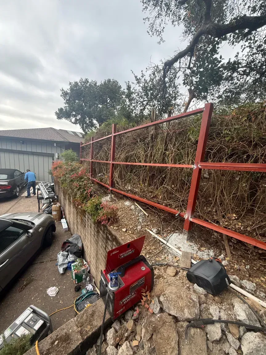A red metal railing being installed atop a stone retaining wall next to a parked car and welding equipment.
