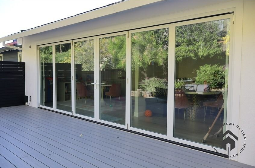 A white-framed bi-fold glass door opening onto a gray wooden deck, showing a patio dining area inside.