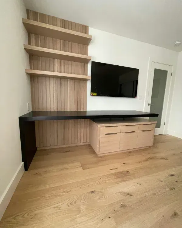 A modern built-in desk unit with light wood cabinetry, open shelving, a black desktop, and a wall-mounted TV.