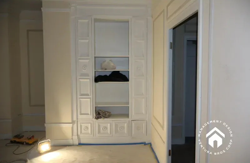 White custom built-in shelving with decorative paneling and molding sits against a wall in a room under renovation.
