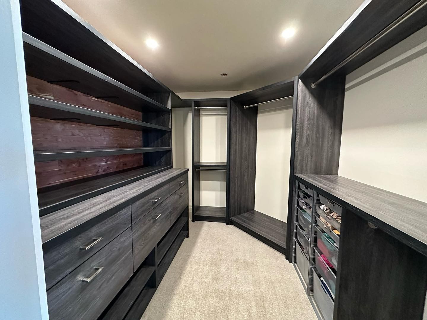 A walk-in closet featuring dark wood cabinets, open shelving, drawers, a hanging rack, and light-colored carpeting.