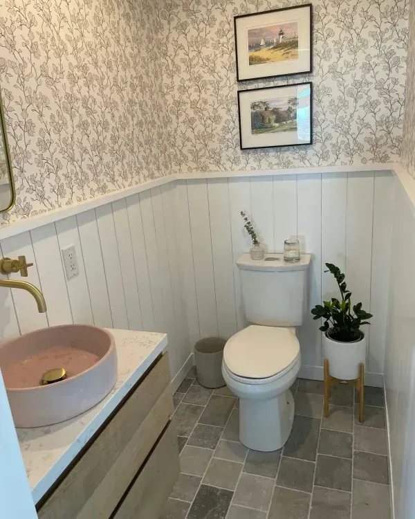 A small bathroom with floral wallpaper, white vertical wall paneling, a pink vessel sink, a toilet, and a potted plant.