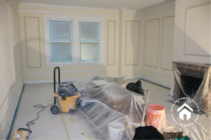 A light-walled room undergoing interior renovation, featuring wainscoting, a fireplace, and construction equipment.