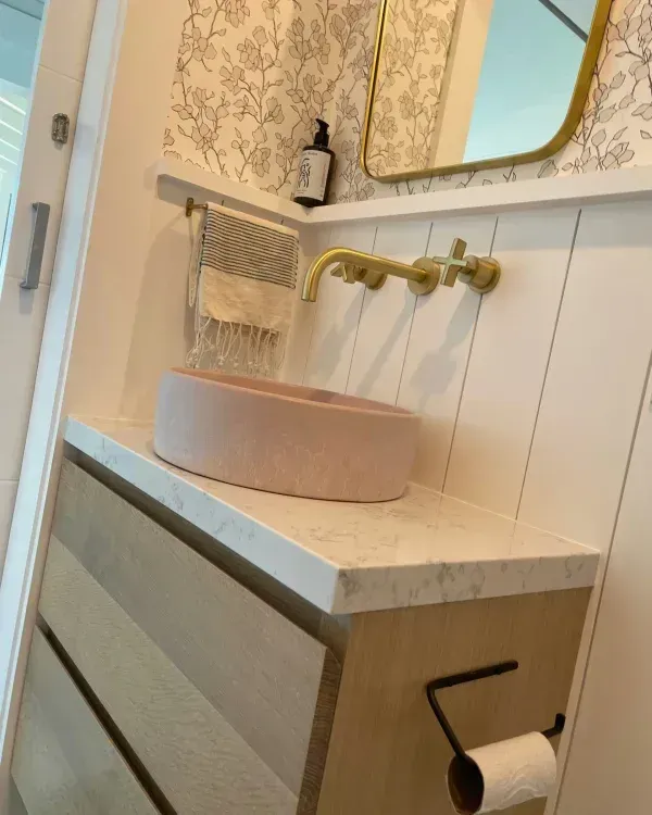 A modern bathroom vanity with a pale pink vessel sink, gold wall-mounted faucet, and light wood cabinetry.