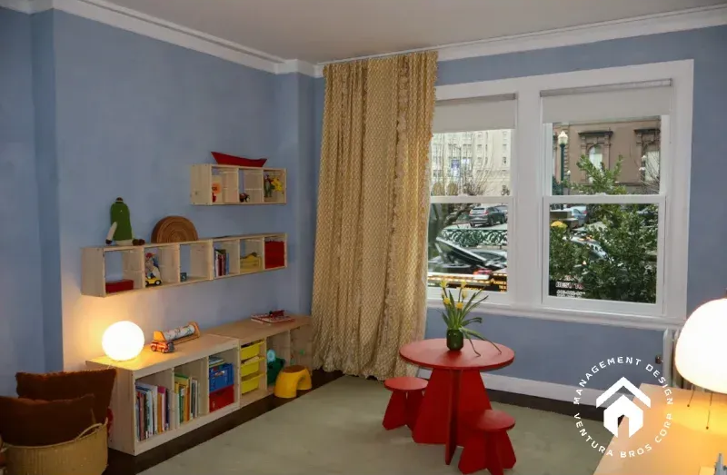 A light blue room featuring a play area with light wood shelves, a red table with two stools, and a large window.
