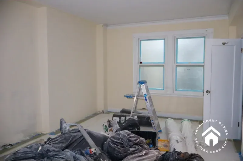 A room under renovation with beige walls, a ladder, construction materials, and a white door near two windows.