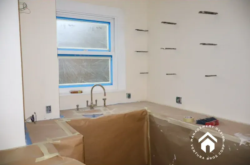 A kitchen under construction, featuring a sink by a window, protective paper covering countertops, and floating shelves.