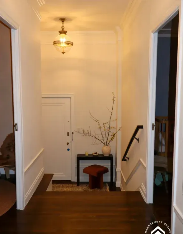 A narrow entryway with wood floors, a white door, a hanging pendant light, and a small console table with branches.