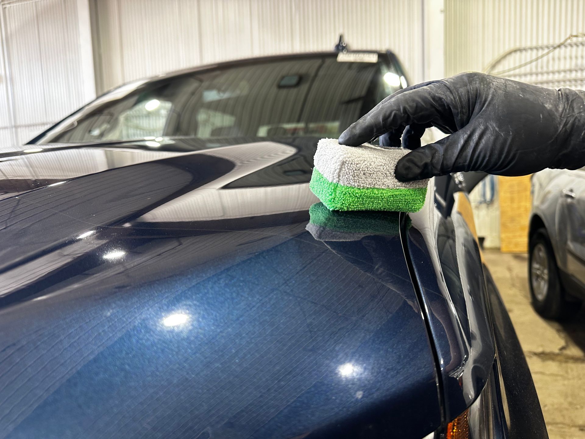 Gloved hand scrubbing a blue car hood with a sponge in a garage