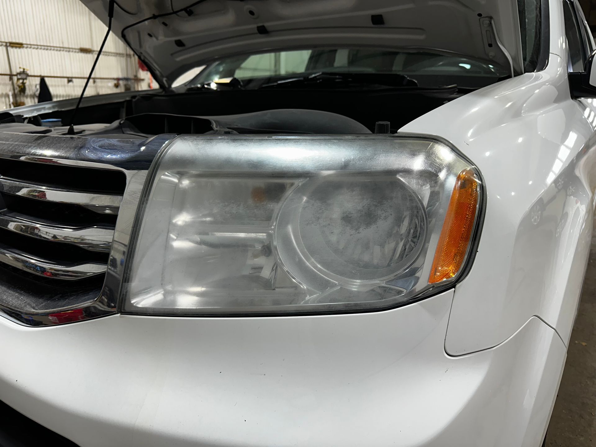 Close-up of a white car front with open hood, grille, and headlight in a garage.