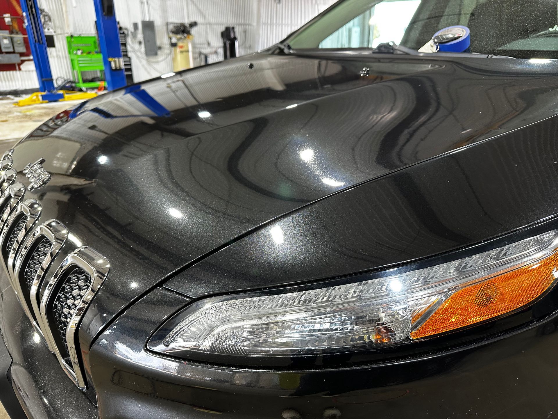 Close-up of a black car hood and headlight in a garage, with blue tape and bright reflections