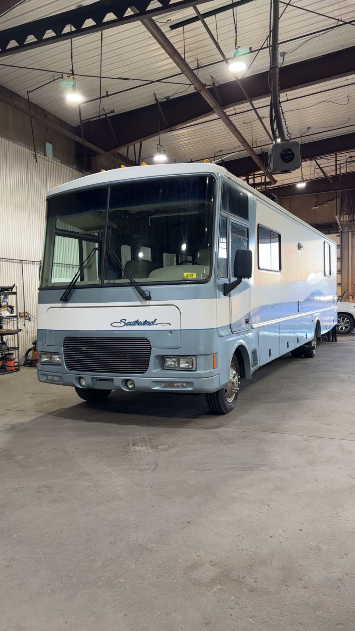 Light blue and white bus parked inside an industrial garage with a high metal ceiling.