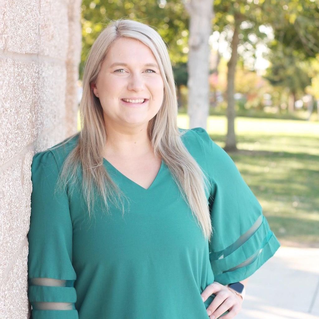 A woman in a green shirt is leaning against a wall and smiling.
