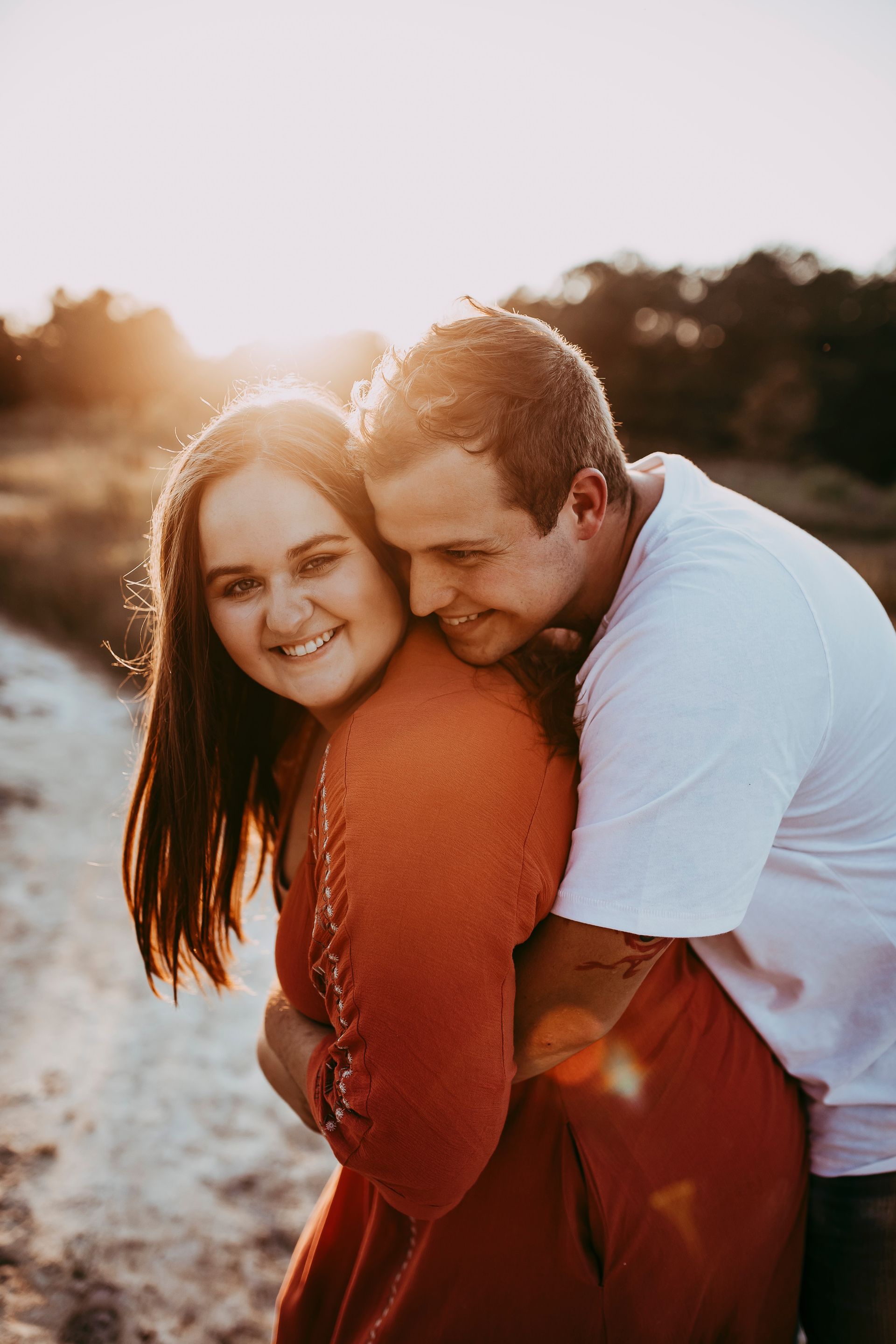 Couple hugging at sunset; woman in red dress smiles, man in white shirt leans in.