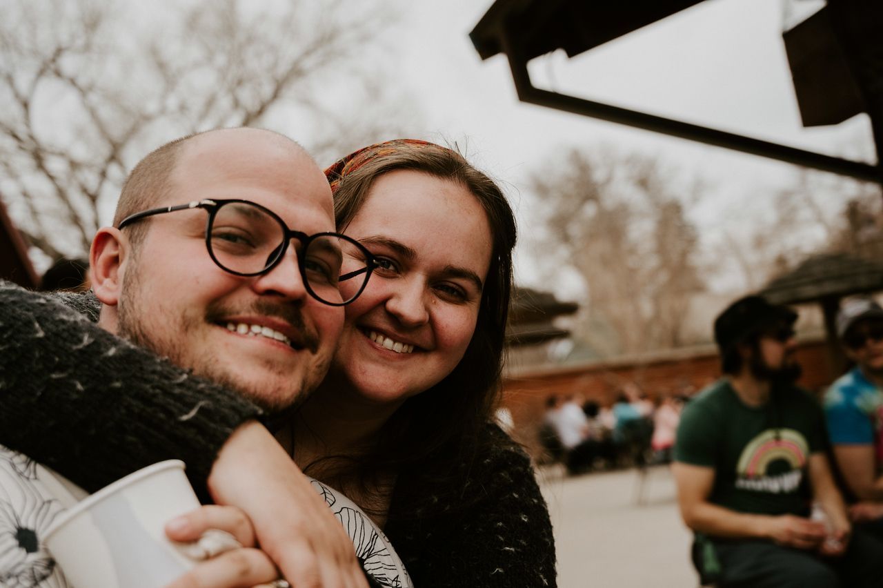 Smiling couple embracing outdoors, with blurred background of a gathering.