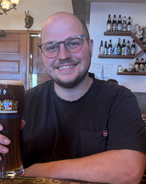 Man with glasses smiling, holding a glass of dark beer in a bar with bottles on shelves.