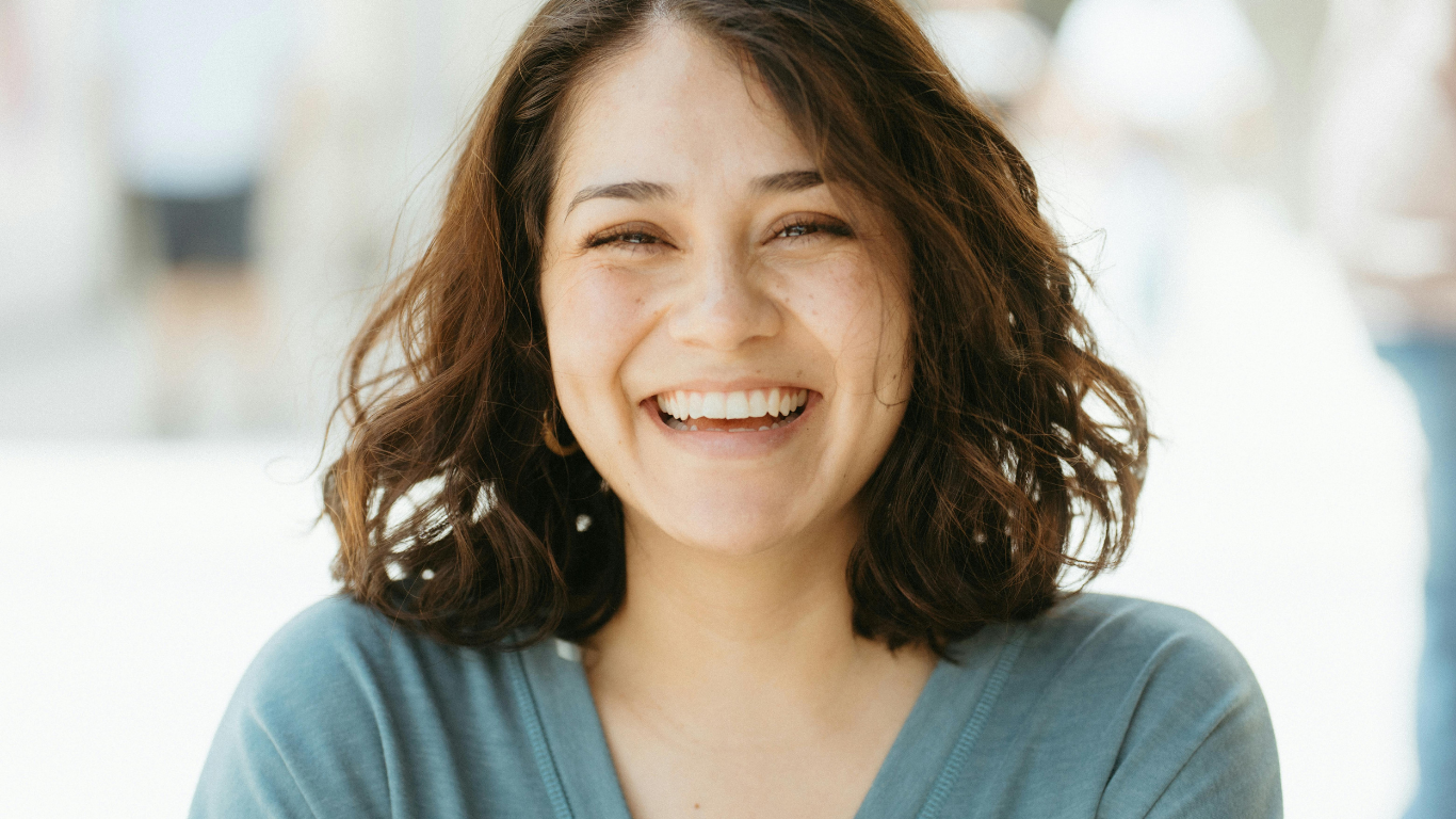 Smiling woman with shoulder-length dark hair in a teal top, standing against a bright blurred background