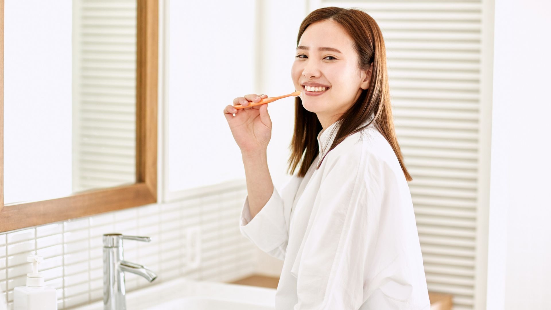 Person brushing teeth in a bright bathroom, smiling beside a sink and mirror.