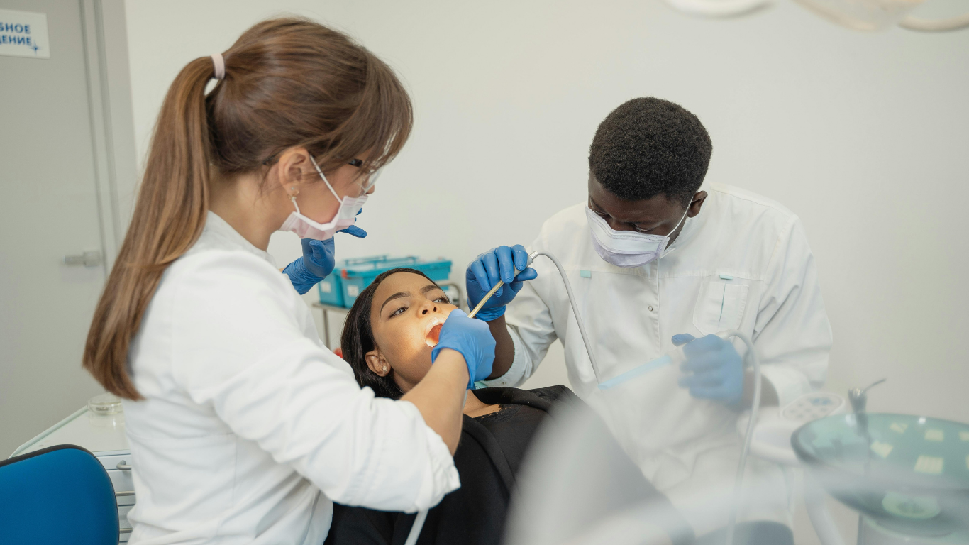 Dental clinicians treat a patient in a bright clinic, using tools and masks during an exam.