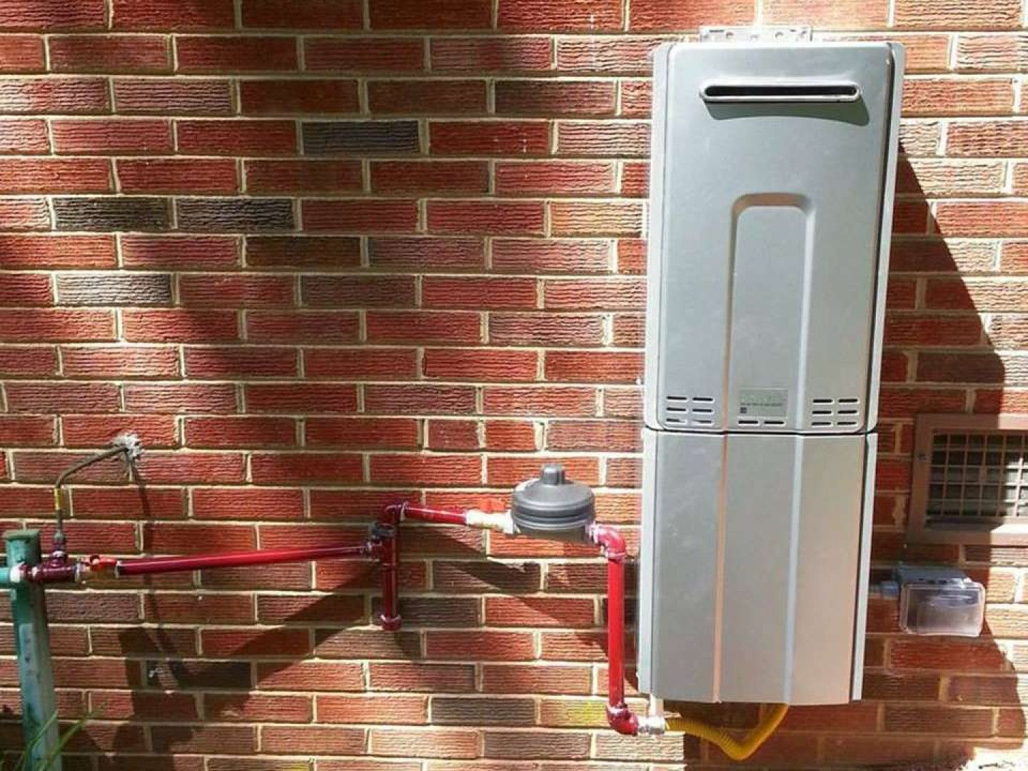 A tankless water heater on a red brick wall, with connected pipes and gas line.