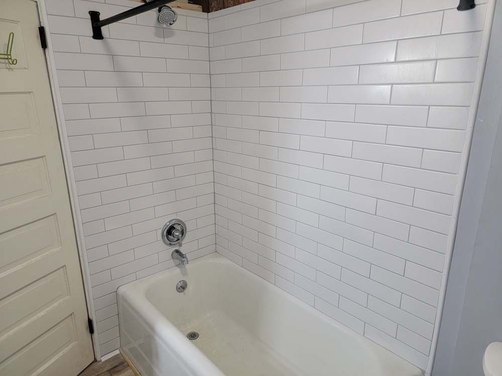 White tiled shower with a bathtub, black shower rod, and silver faucet.