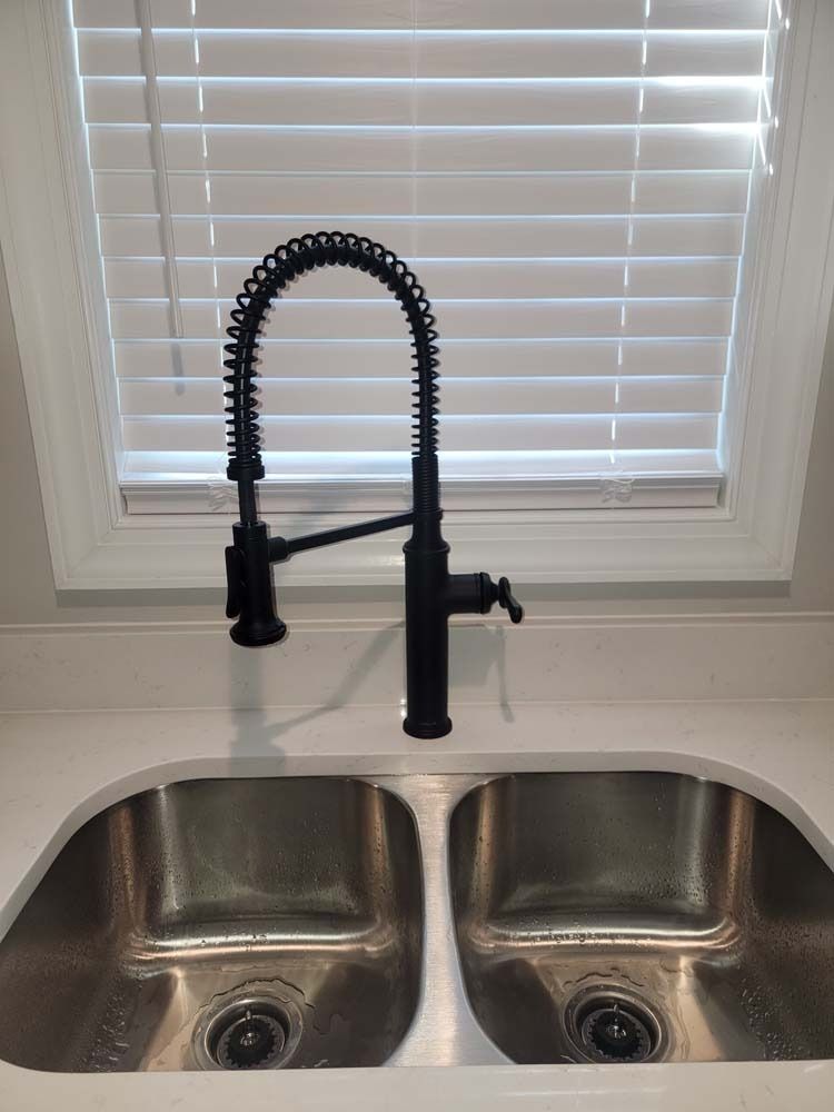 Black kitchen faucet over a stainless steel double sink, white countertop, window with blinds.